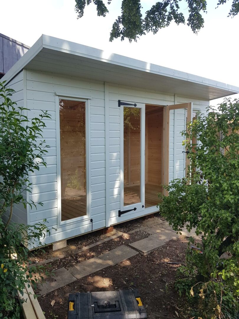 White-painted garden studio with tall glass doors and windows, flat roof, and timber interior, surrounded by plants