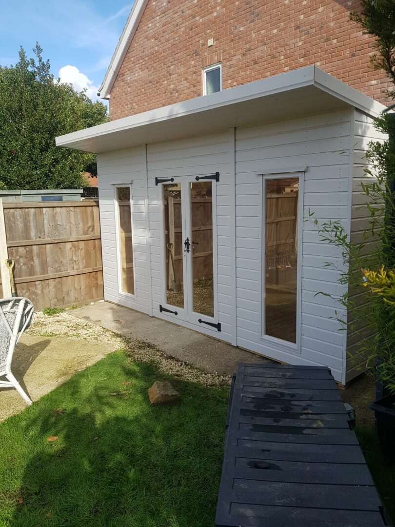 White-painted garden office with flat roof, double doors, and tall windows set beside a brick house and lawn