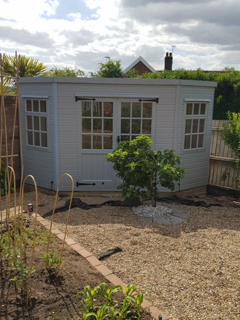 Grey-painted summer house with double Georgian-style doors and windows, set in a gravel garden with raised beds and tree