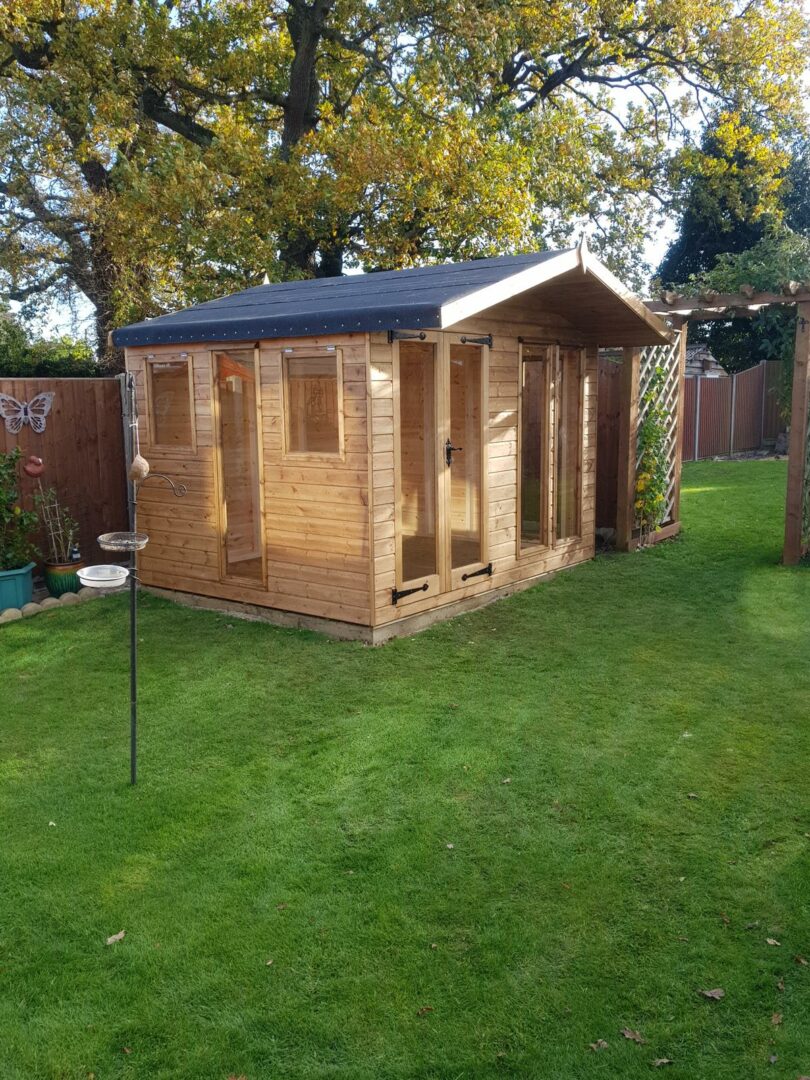Wooden garden office studio with windows and double doors, surrounded by green lawn and trees in autumn