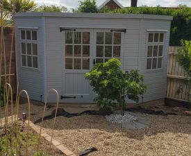 Grey-painted summer house with double Georgian-style doors and windows, set in a gravel garden with raised beds and tree